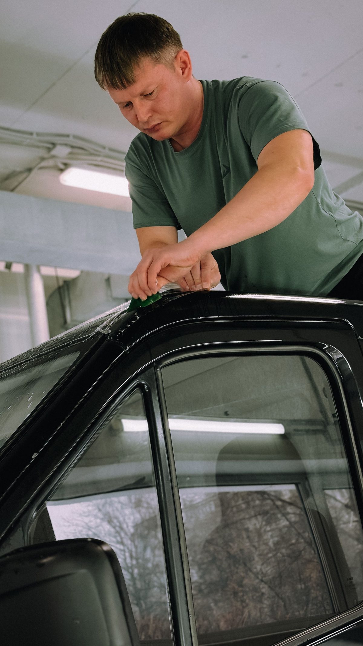 Installer in an olive shop tee squeegeeing paint protection film onto a black car.